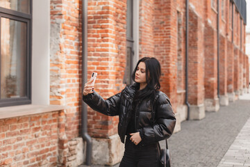 Fototapeta na wymiar Pensive brunette woman make selfie posing on street brick building background. Outdoor shot of happy hippie lady make video call, smiling. Girl wear black puffer jacket raises her hand hold camera.