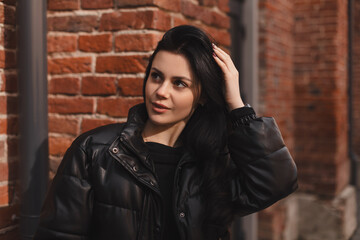 Trendy girl with brunette hairstyle posing outside. Woman in leather puffer jacket fix hair,...