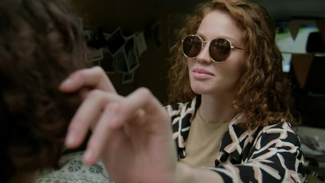Stylish girl in sunglasses sitting in camper van in front of her boyfriend, stroking his hair and smiling to him while enjoying spending time together on trip