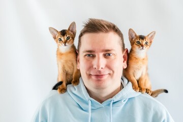 Smiling funny man in blue hoodie with twin Abyssinian cats, small newborn kittens sitting on...