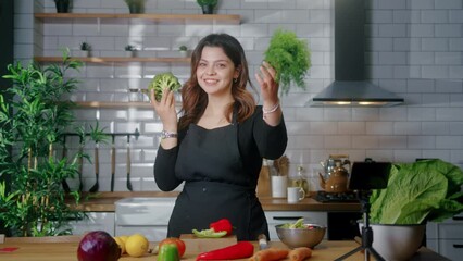 Young vlogger woman in black cooking apron looking at camera showing green vegetables to camera smiling. Slow motion
