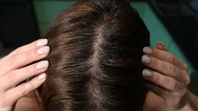 a woman touches her hair, looking at early gray hair. Parting of dark brown female hair with gray roots. close-up