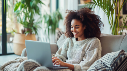 Smiling woman using a laptop while sitting on a sofa at home showcasing a cozy and modern lifestyle, suitable for content about remote work or leisure. - obrazy, fototapety, plakaty