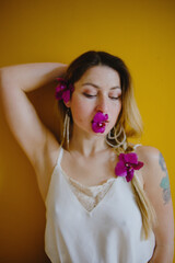 Portrait of a blonde girl against a yellow wall. An orchid flower in your mouth and in your hair