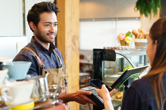 Male cafe employees of various nationalities holding  credit card acceptor showing QR code smiled. young brunette customer holding mobile phone scanning pay coffee bakery products cafe small business.