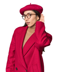 Stylish young Caucasian woman in red trench coat and beret on studio background trying to listening a gossip.