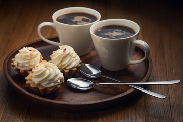 Two white cups with black coffee, three cream-topped pastries on a dark wooden tray, two spoons