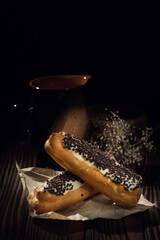 Two eclairs with cream and chocolate chips beside a black mug on a dark wooden surface, softly lit with a rustic backdrop.
