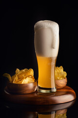 Frothy beer with overflowing head beside bowls of chips on a wooden surface against a dark backdrop.