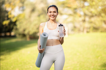 A joyful active woman holds a yoga mat and water bottle, radiating health