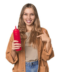 Blonde woman holding ketchup bottle in studio setting person pointing by hand to a shirt copy...