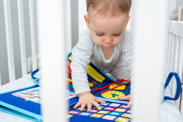 Baby playing with montessori busy book sitting in crib. Concept of smart books and modern toys