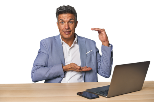 Elegant businessman at desk with laptop shocked and amazed holding a copy space between hands.