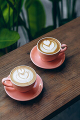 vertical shot of two coral-colored coffee mugs with intricate latte art, resting on a rustic wooden...