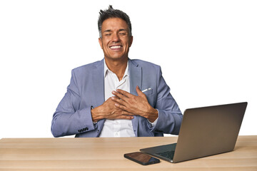 Elegant businessman at desk with laptop laughing keeping hands on heart, concept of happiness.