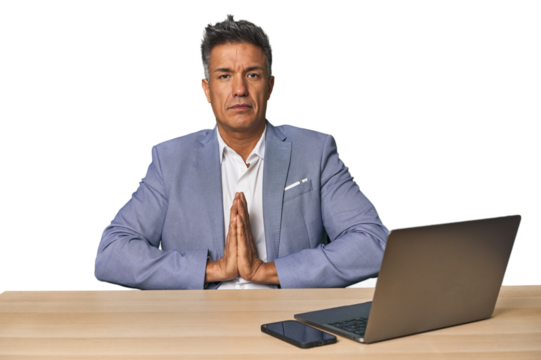 Elegant businessman at desk with laptop praying, showing devotion, religious person looking for divine inspiration.