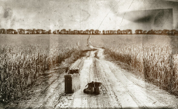 Ankle leather boots lie in the middle of a dirt road in the wheat fields. A film camera and an antique cardboard suitcase stand next to it. An old scratched and smudged black-and-white photo effect