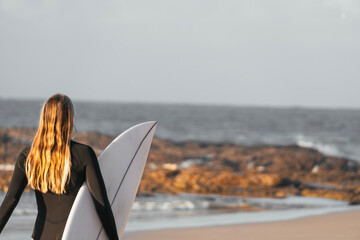 Blonde surfer girl walking along the beach shore with her surfboard alone