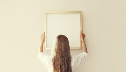 Woman decorating interior, hanging white blank photo frame, wooden picture on wall in new house