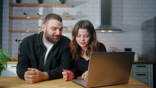 Smiling young couple sit in the kitchen woman enter credit card number on laptop for shopping online, makes secure easy distant electronic payment
