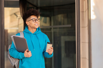 Smiling student guy holding takeaway coffee, laptop and workbooks outside