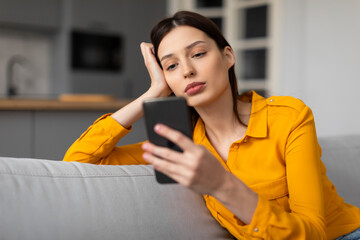 Pensive woman with phone looking sad and thoughtful on sofa