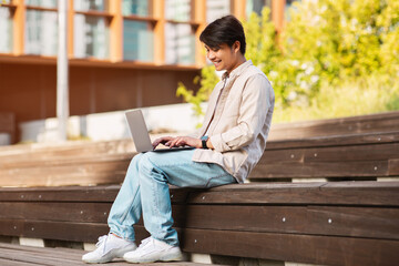 Asian guy sitting on bench at urban area, using laptop