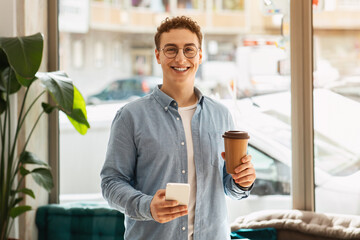 Smiling young man with curly hair and glasses holding a smartphone in one hand and a takeaway...