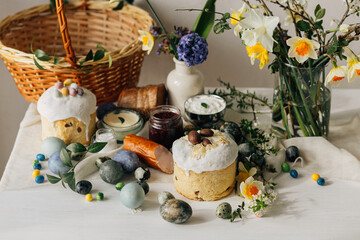 Stylish Easter eggs, bread and basket with spring flowers on rustic table. Happy Easter! Traditional easter holiday food for blessing. Modern natural dye eggs, tasty ham, bread, butter, beets