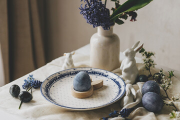 Naklejka premium Happy Easter! Stylish easter egg in bunny figurine on vintage plate and spring flowers on linen, rustic table setting. Natural painted blue eggs and hyacinth blooms. Modern minimal still life
