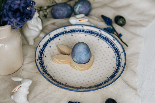 Happy Easter! Stylish easter egg in bunny figurine on vintage plate and spring flowers on linen, rustic table setting. Natural painted blue eggs and hyacinth blooms. Modern minimal still life