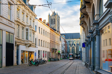 A tram on tracks comes through Sint-Niklaasstraat, or Saint Nicholas Street, with the tower and nave of the Saint Nicholas Cathedral in view behind, in Ghent.