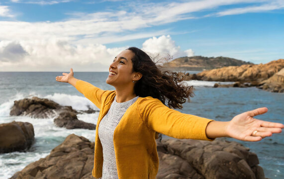 A woman is standing on a rocky beach, smiling and waving her arms in the air