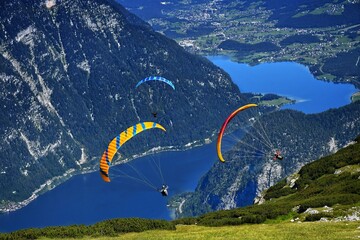 Two athletes are paragliding over a lake in Austria surrounded by green forest and blue sky above...