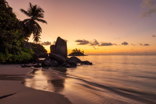 Orange hues of a beautiful sunrise on a romantic beach with palm trees and granite rocks on a tropical island in Seychelles, Africa