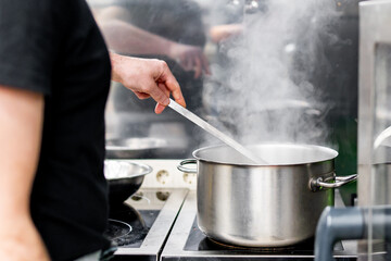 Professional chef in black attire stirring a large steaming pot on a stove in a commercial kitchen, with various kitchen utensils in the background