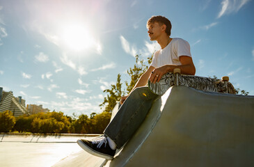 Young male skateboarder holding skateboard sitting on ramp in skate park 