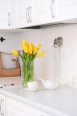 Home kitchen interior.Vase with bouquet of yellow tulips, glass of tea and teapot on white worktop of kitchen cupboard. Spring consept.