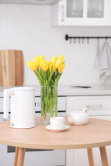 Home kitchen interior.Vase with bouquet of yellow tulips, electric teapot, glass of tea on wooden table. Spring consept.