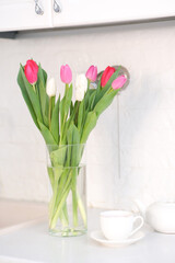 Home kitchen interior.Vase with bouquet of pink and white tulips, glass of tea, teapot on white worktop of kitchen cupboard. Spring consept.