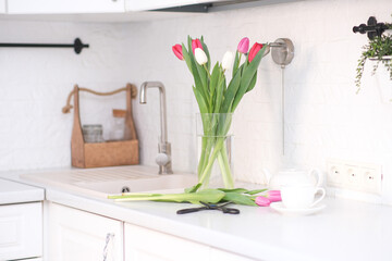 Home kitchen interior.Vase with bouquet of pink and white tulips, glass of tea, sissors, teapot on...