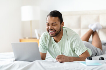 Happy african american guy watching webinar, bedroom interior