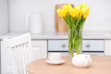 Home kitchen interior.Vase with bouquet of yellow tulips, electric teapot, glass of tea on wooden table. Spring consept.
