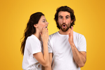 Surprised bearded man with wide eyes and an open mouth holds his hands up while a woman whispers into his ear