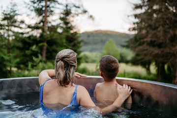 Mother with her little son enjoying view at nature while bathing in wooden barrel hot tub in the terrace of the cottage.