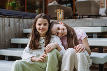 Two sisters sitting on stairs in front of house, embracing. Sisterly love and siblings relationship...