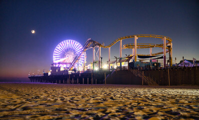 Eindrücklich beleuchtetes Riesenrad und Achterbahn am Santa Monica Pier am Abend mit Halbmond im...