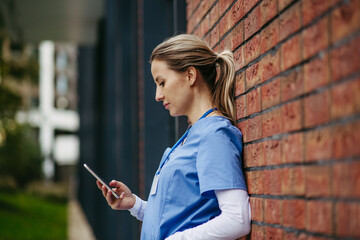 Nurse, doctor in uniform taking break, standing in front hospital building, scrolling on...