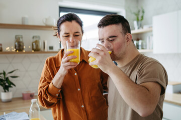 Fototapeta premium Portrait of young man with Down syndrome with his mother at home, toasting, drinking juice. Morning routine for man with Down syndrome.