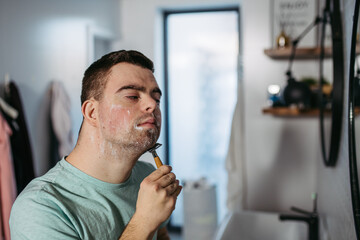 Young man with down syndrome learning how to shave, holding razor in hand, looking at mirror.